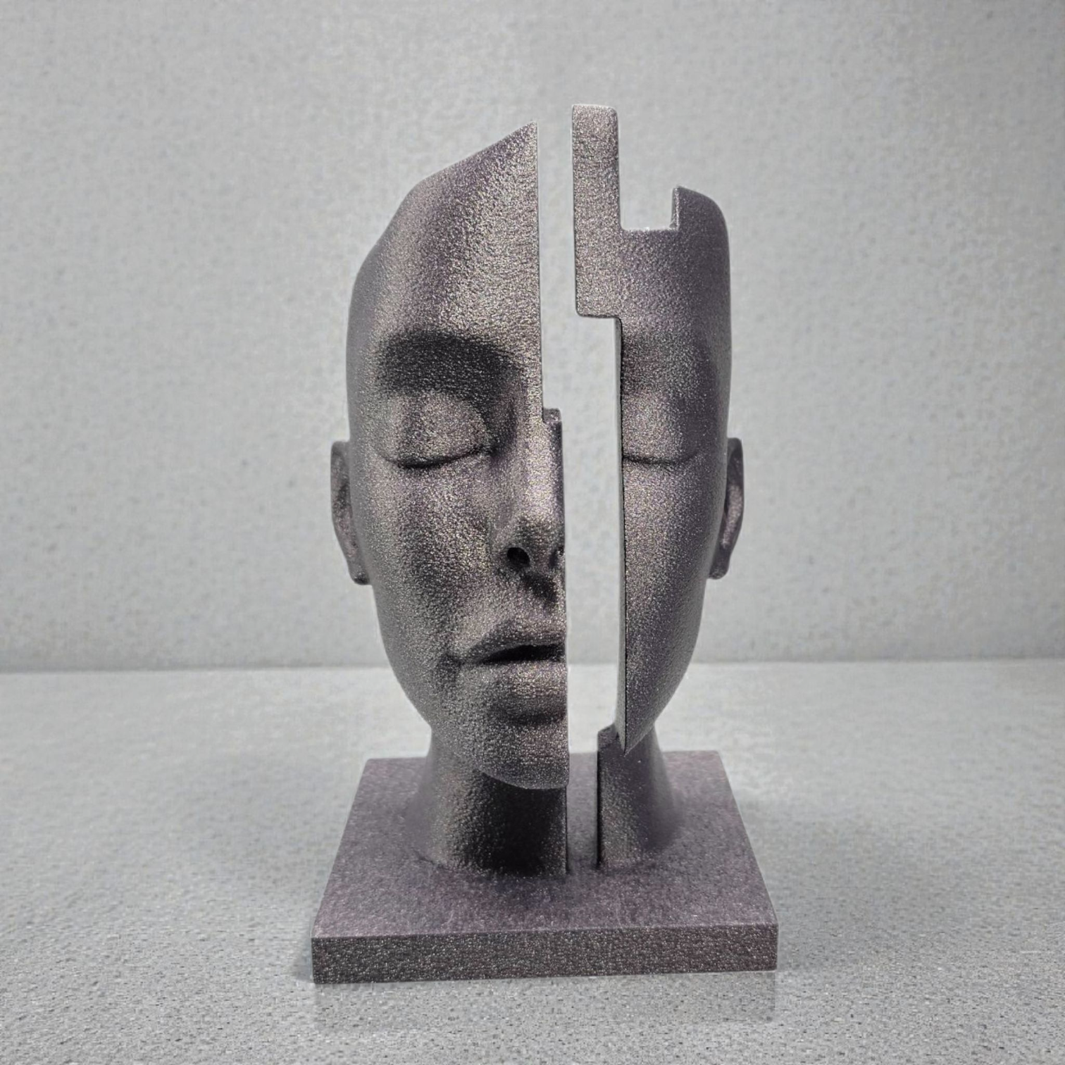 Zilveren sculptuur van een verticaal gesplitst menselijk gezicht met gesloten ogen en een vierkante sokkel, geplaatst tegen een gespikkelde achtergrond.
