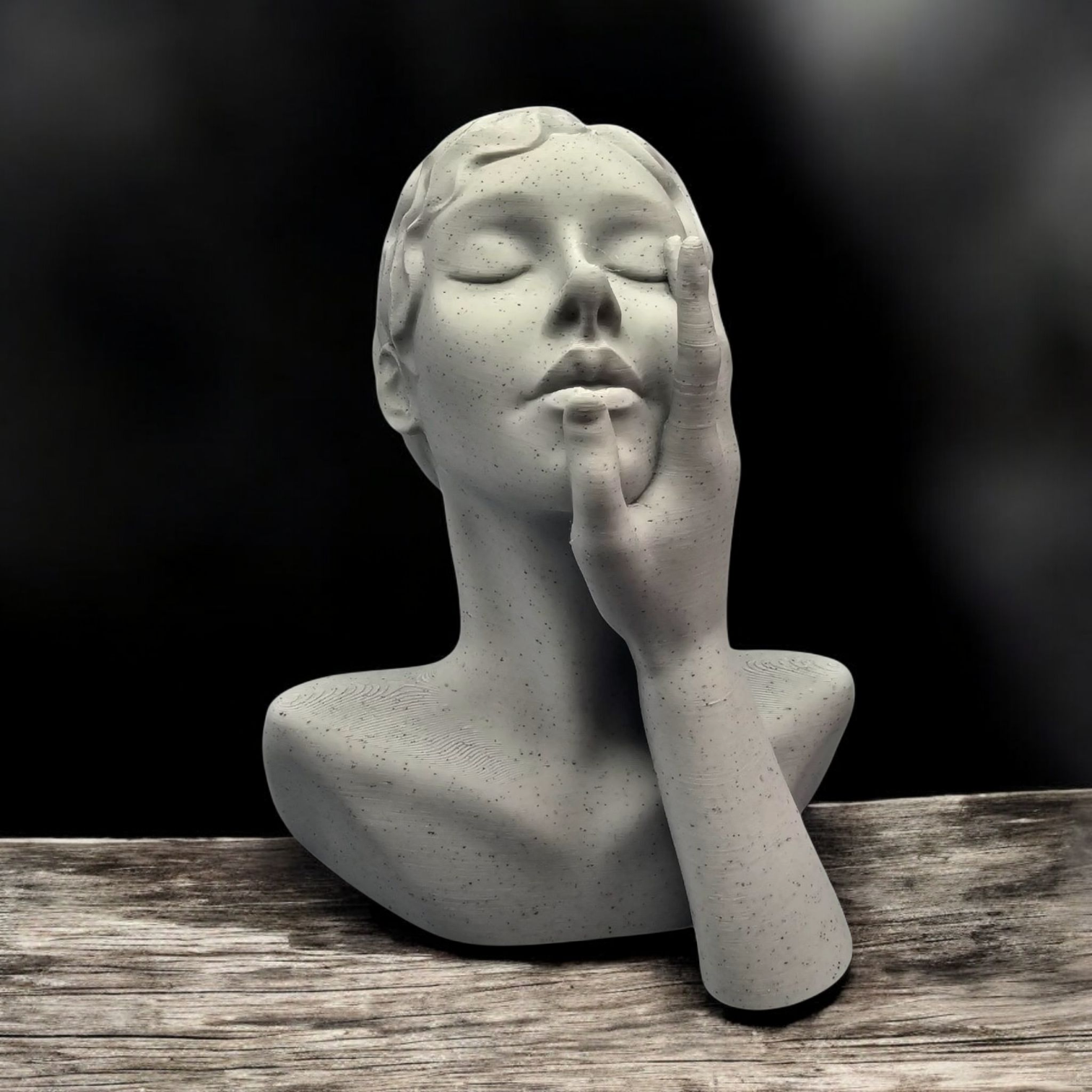 Stenen buste van een vrouw met gesloten ogen, haar hand tegen haar gezicht, geplaatst op een houten ondergrond.