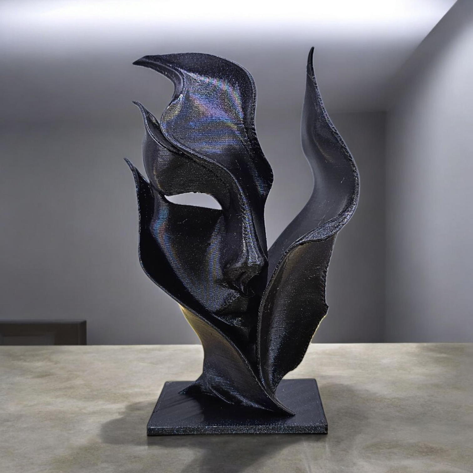 Zwarte abstracte sculptuur, die doet denken aan een vloeiende stof of vlammen, staat op een vierkante sokkel op een tafel.