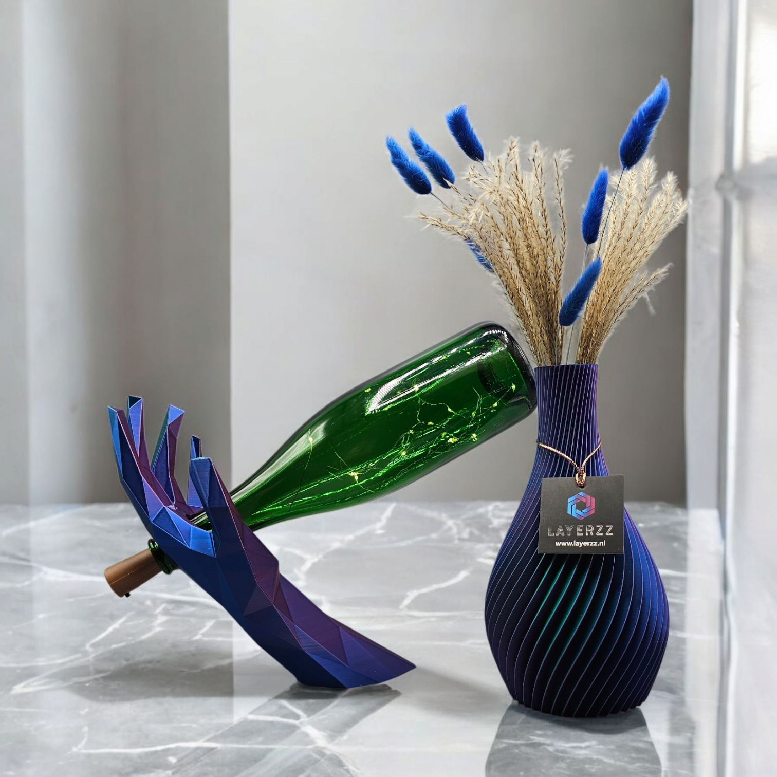 Wijnfles in een blauwe, handvormige standaard; vaas met blauwe gedroogde bloemen, op een grijze ondergrond, naast het raam.