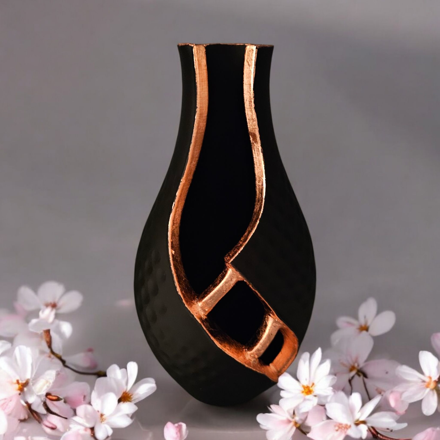Zwarte vaas met koperkleurige accenten, geplaatst voor wazige witte en roze bloemen.