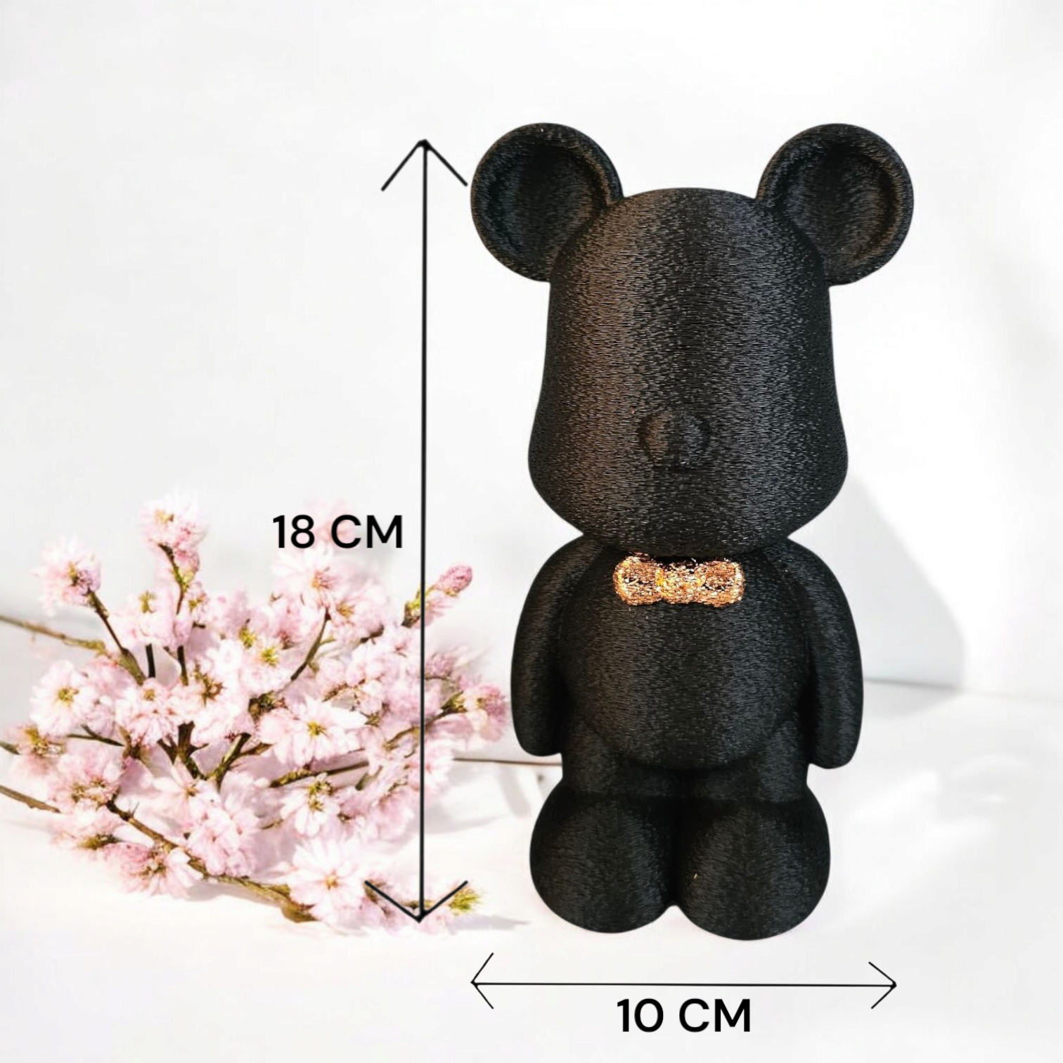 Zwarte teddybeer met gouden strik, 18 cm hoog, naast roze bloemen.