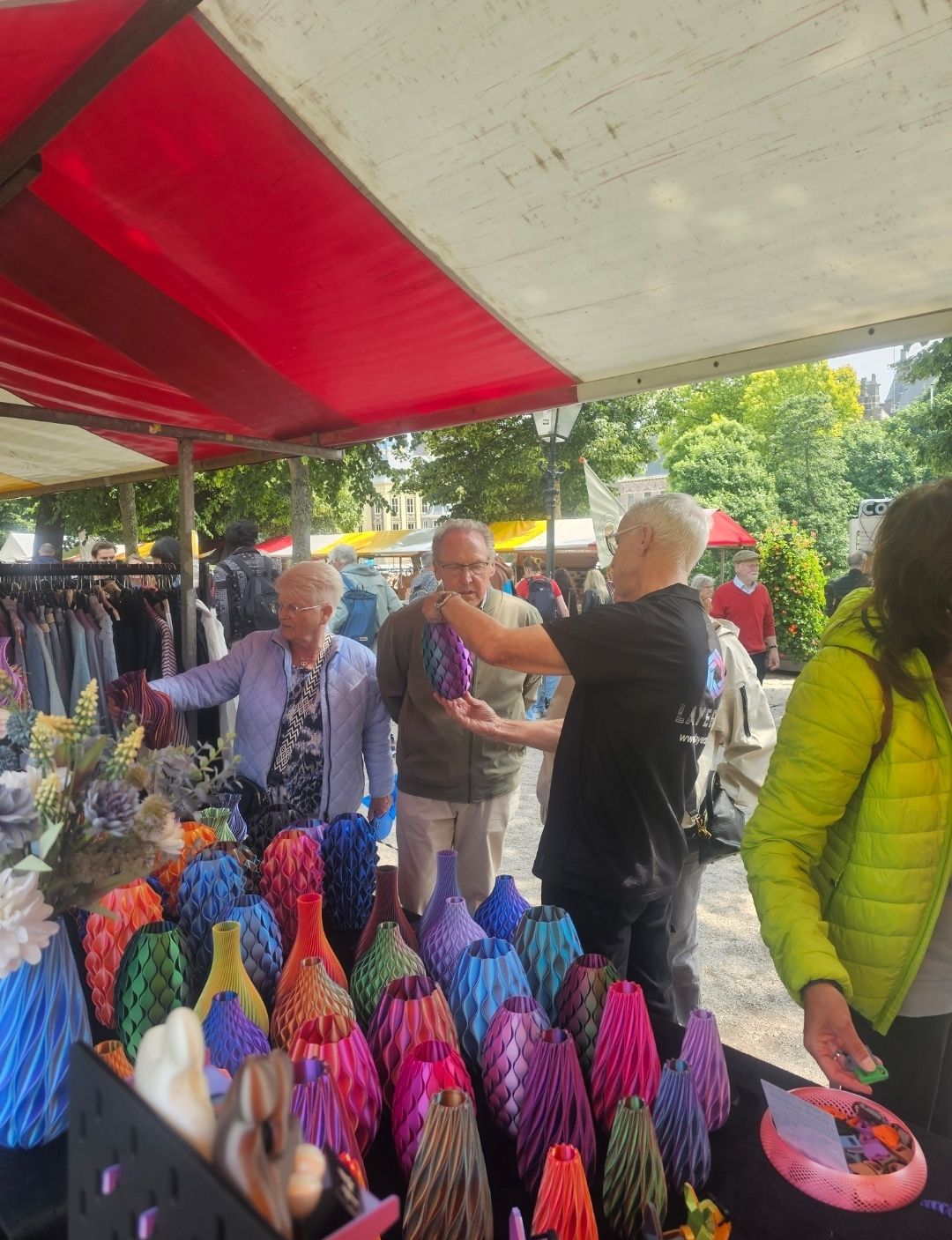 Mensen bekijken kleurrijke vazen op een openluchtmarkt onder een rode luifel.