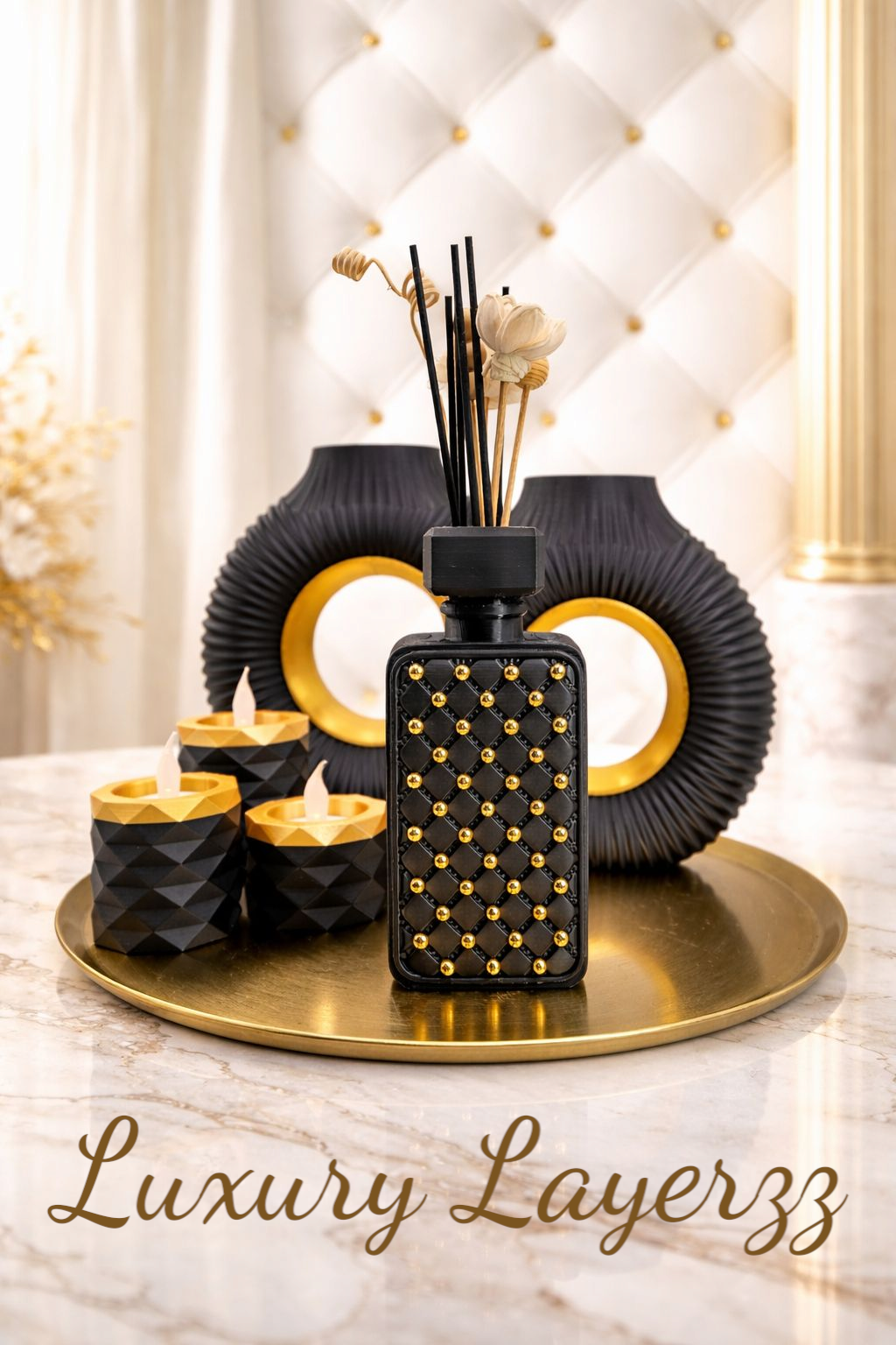 Decoratieve woonaccessoires op een gouden dienblad: geurdiffuser, kaarsen, zwarte vazen, tegen een witte, gewatteerde achtergrond.