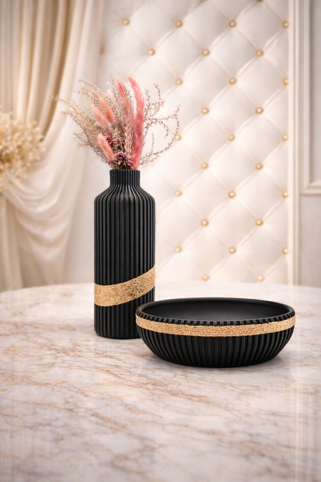 Zwarte vaas en schaal met gouden accenten op een marmeren tafel, gedroogde roze en beige bloemen, tegen een crèmekleurige muur met ruitjespatroon.