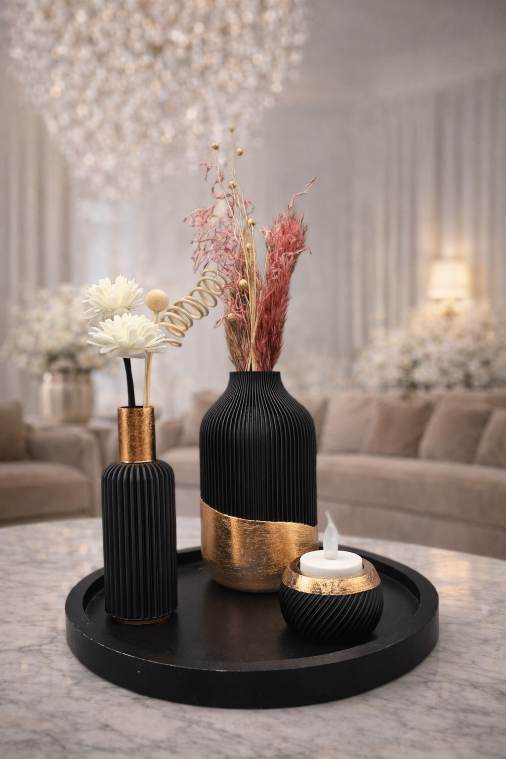 Een decoratieve tafelsetting: zwarte en gouden vazen ​​met gedroogde bloemen en een kaars op een rond dienblad in een woonkamer.
