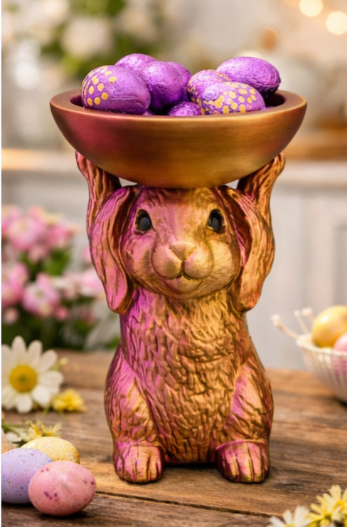 Een gouden konijnenbeeldje met een kom paarse paaseieren staat op een houten tafel buiten.