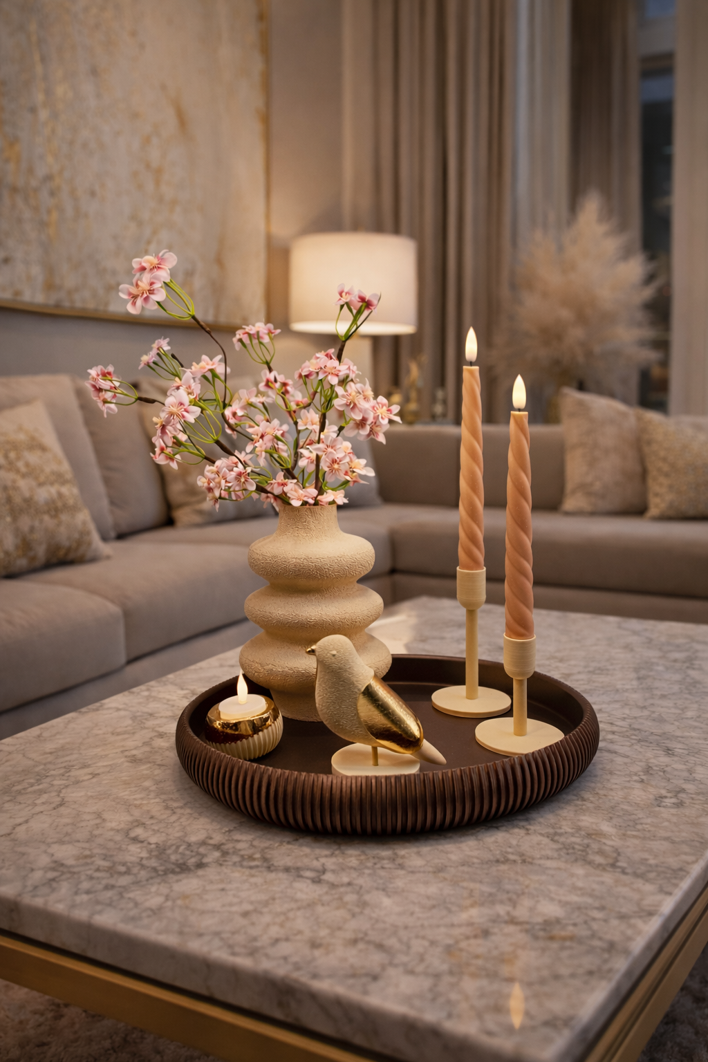 Op een ronde schaal op een marmeren tafel staan ​​een vaas met een bijzondere textuur gevuld met roze bloesem, twee spiraalvormige kaarsen, een vogelornament en nog een kaars.