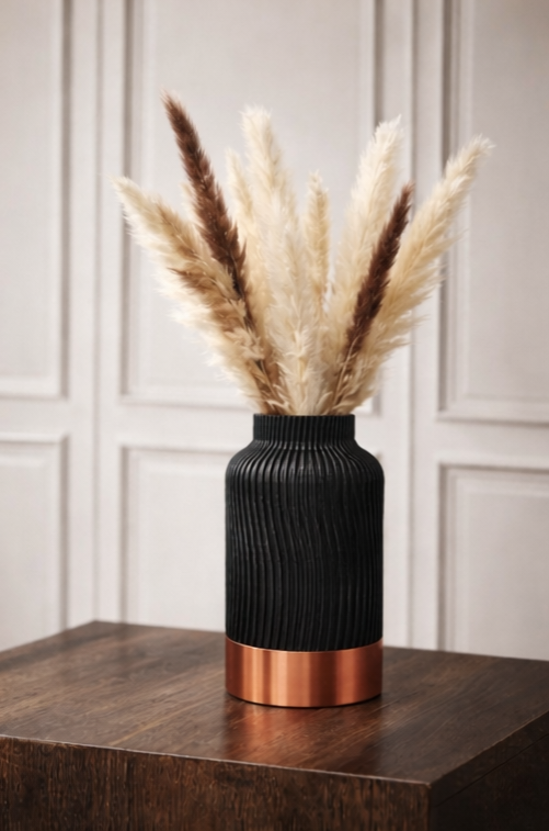 Zwarte vaas met koperen voet, gevuld met pampasgras, op een houten ondergrond.