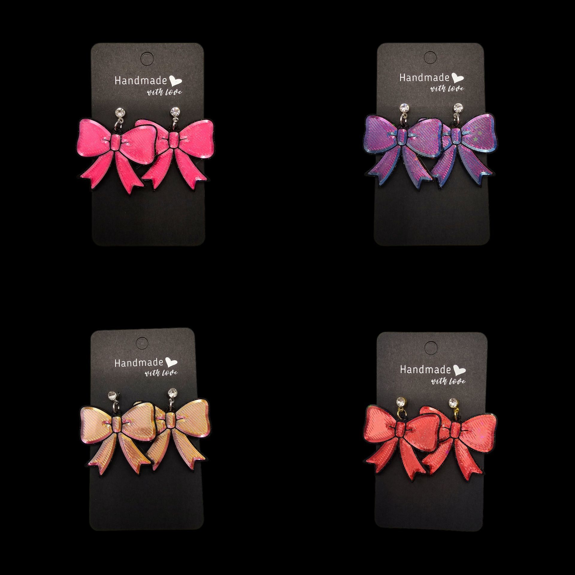 Vier paar strikvormige oorbellen op zwarte kaartjes: roze, paars, goud en rood.