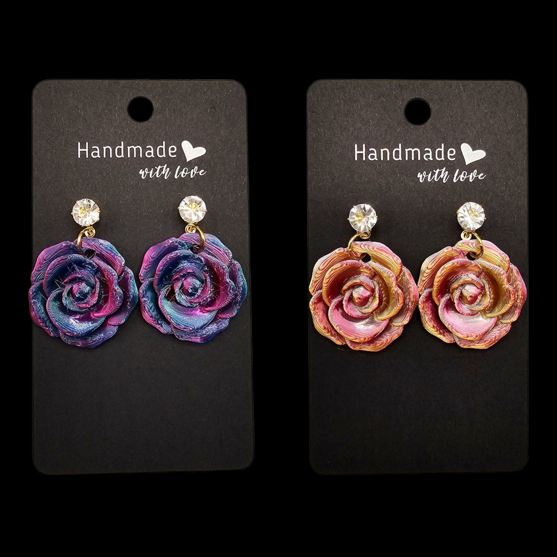 Twee paar handgemaakte rozenoorbellen op zwarte presentatiekaartjes: een paar paars/blauw, het andere roze/goud.