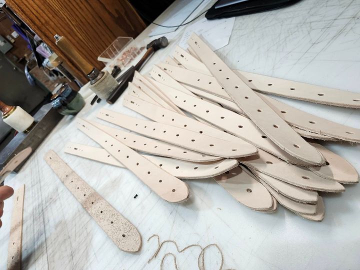 A pile of light-colored, punched leather strips rests on a workbench beside leatherworking tools.