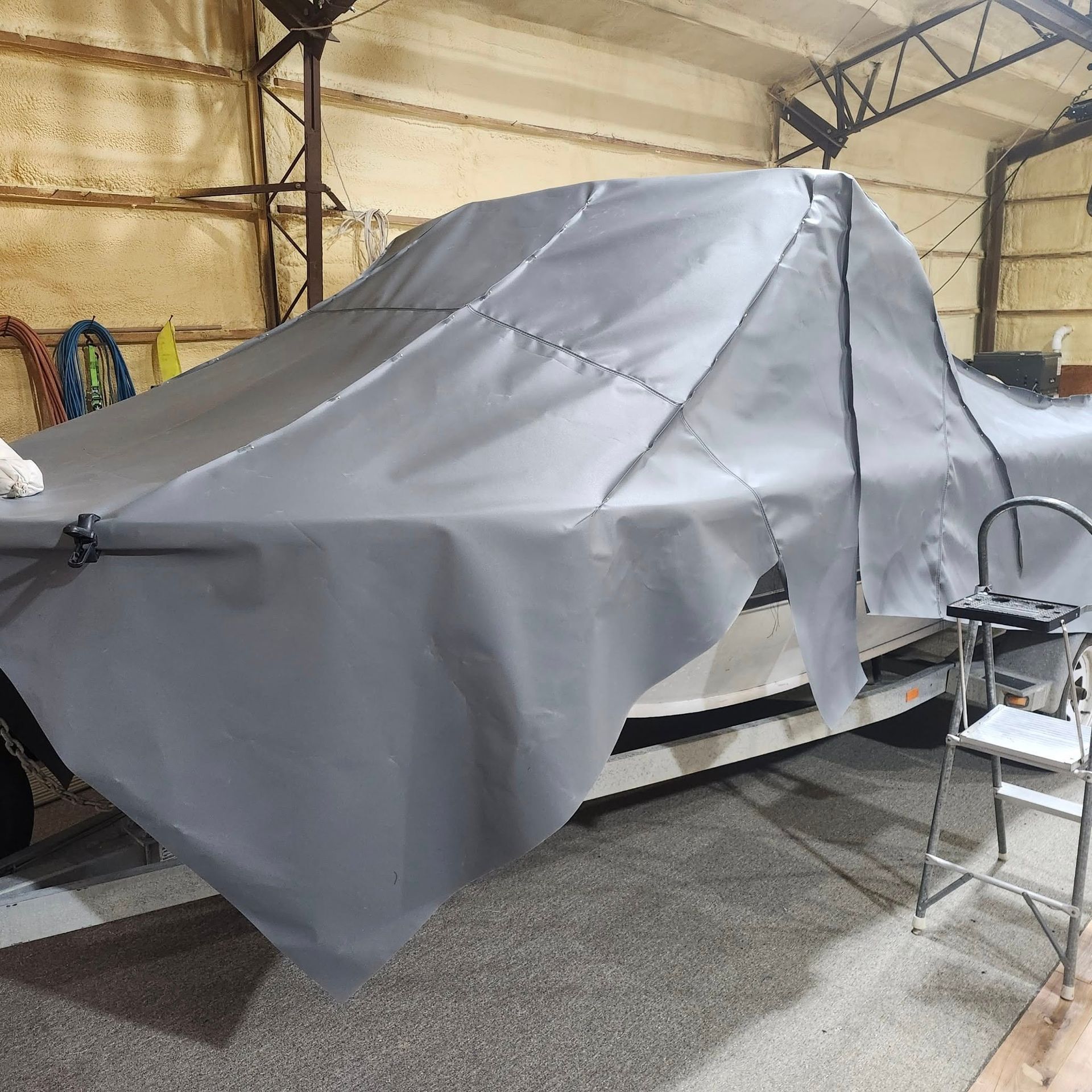 A grey boat cover draped over a boat sitting on a trailer in a garage, with a step ladder nearby.