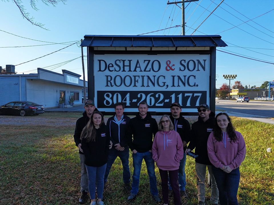 A group of people standing in front of a sign for deshazo & son roofing inc.