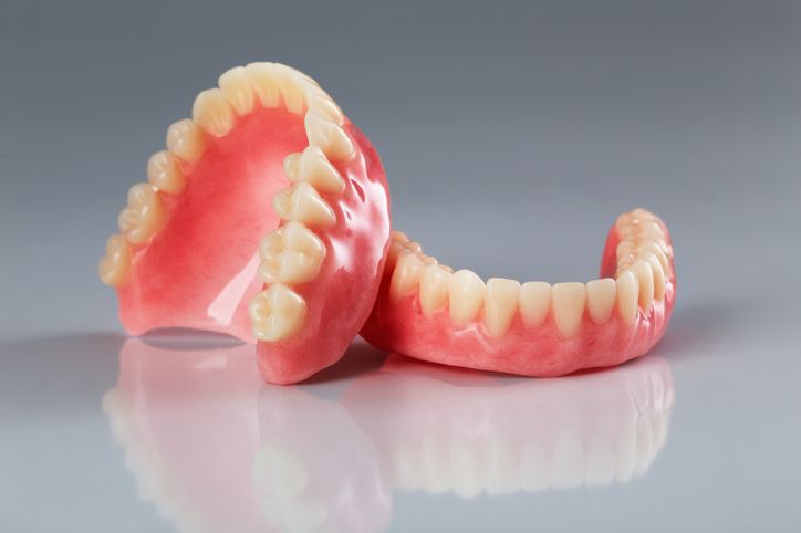 Full dentures, upper and lower, pink gum and white teeth, resting on a reflective surface.