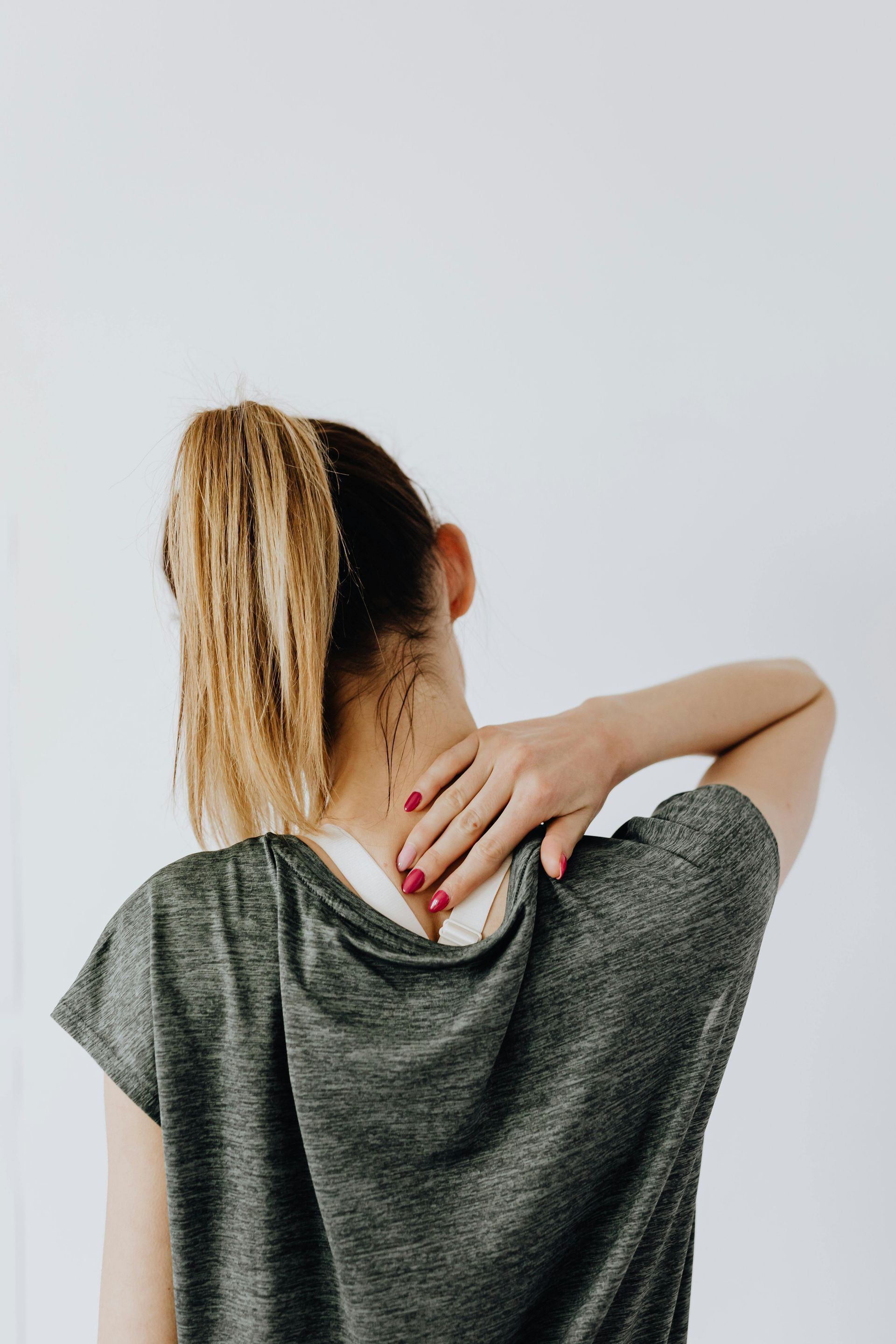 Person with ponytail rubbing neck and shoulder, wearing a gray shirt against a plain light background