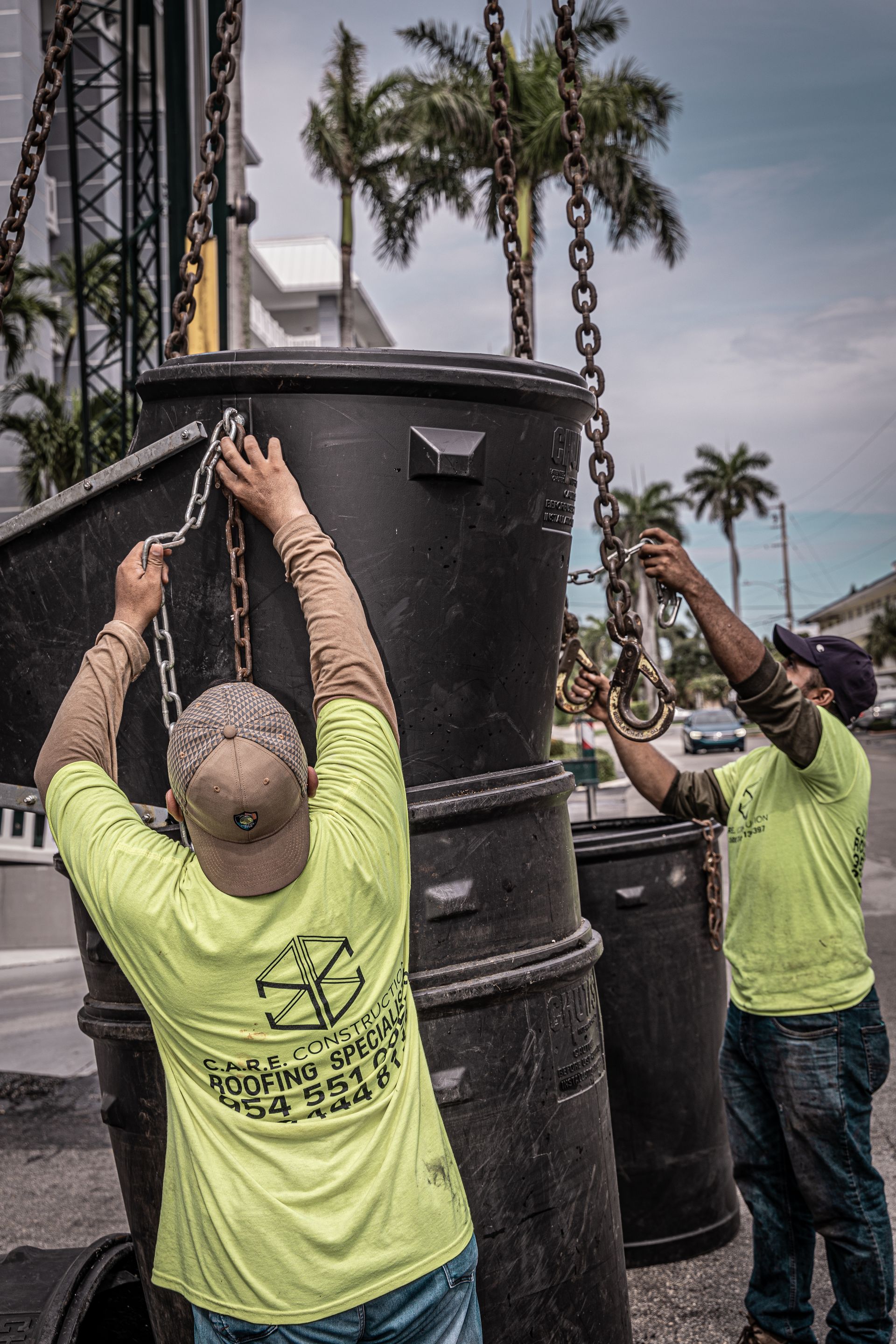 Two men wearing neon yellow shirts are lifting a large trash can