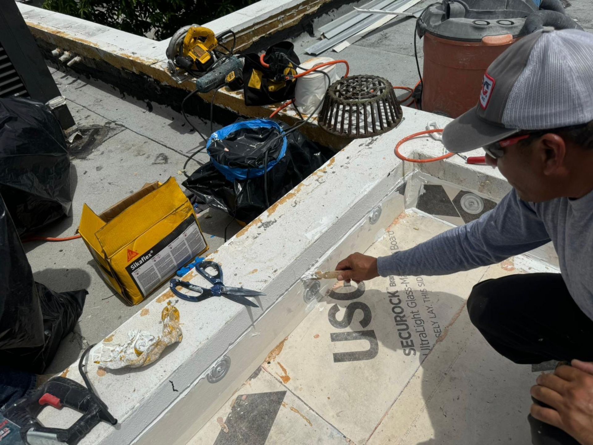 A man is kneeling down on a roof working on a piece of plywood.