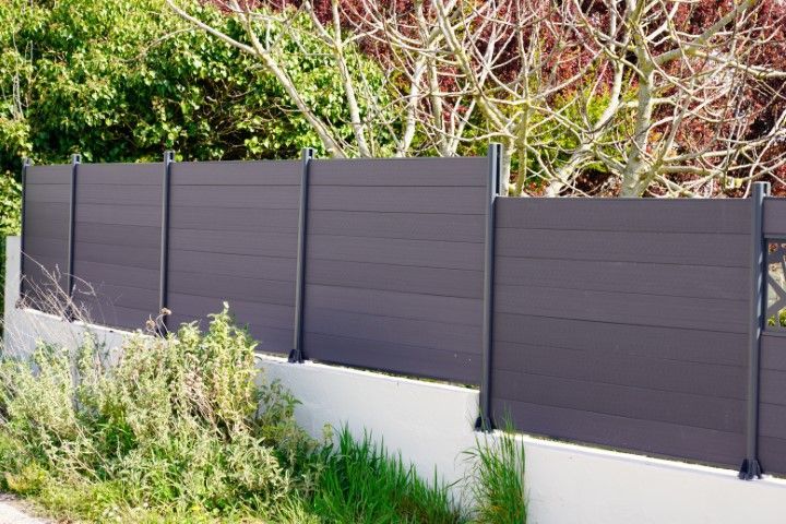 Gray horizontal plank fence on a white wall with metal posts, green foliage in the background.