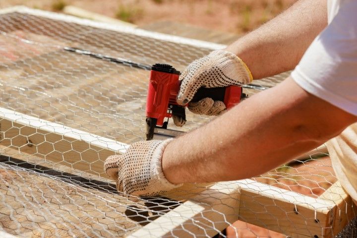 Person wearing gloves stapling chicken wire to a wooden frame with a red staple gun.
