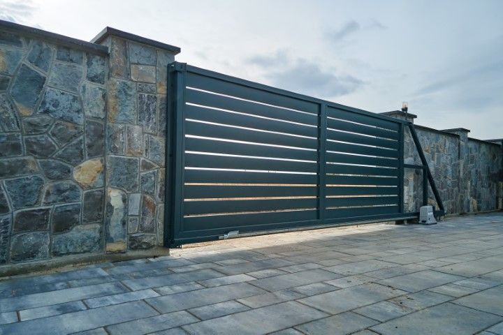 Gray sliding metal gate with horizontal slats, stone wall, and paved driveway.