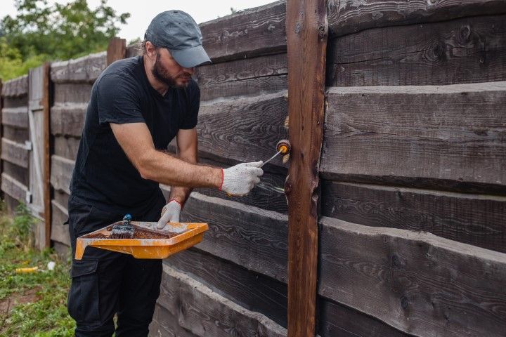 Man painting a wooden fence with a brush, holding a paint tray outside.