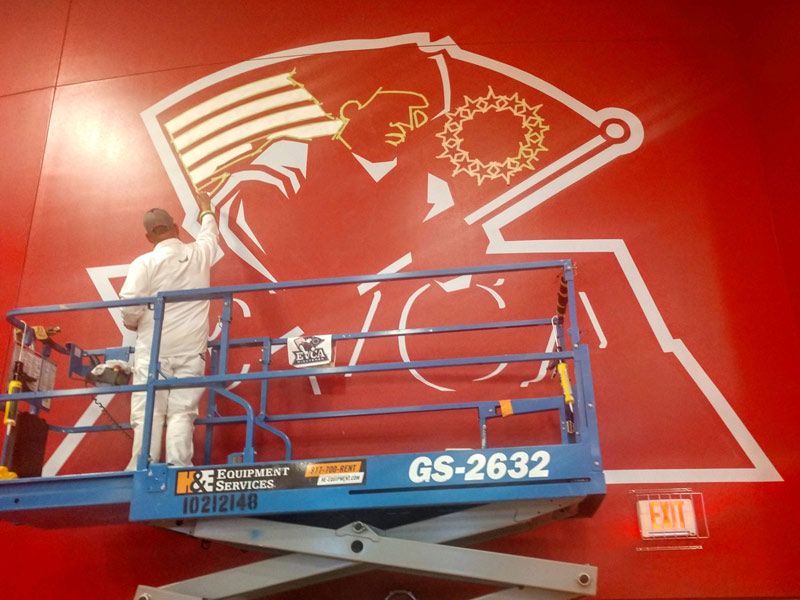 A man is painting a picture on a red wall on a scissor lift.