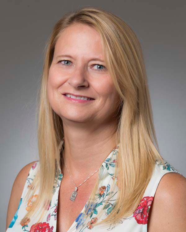 A woman with long blonde hair and a floral shirt is smiling for the camera.