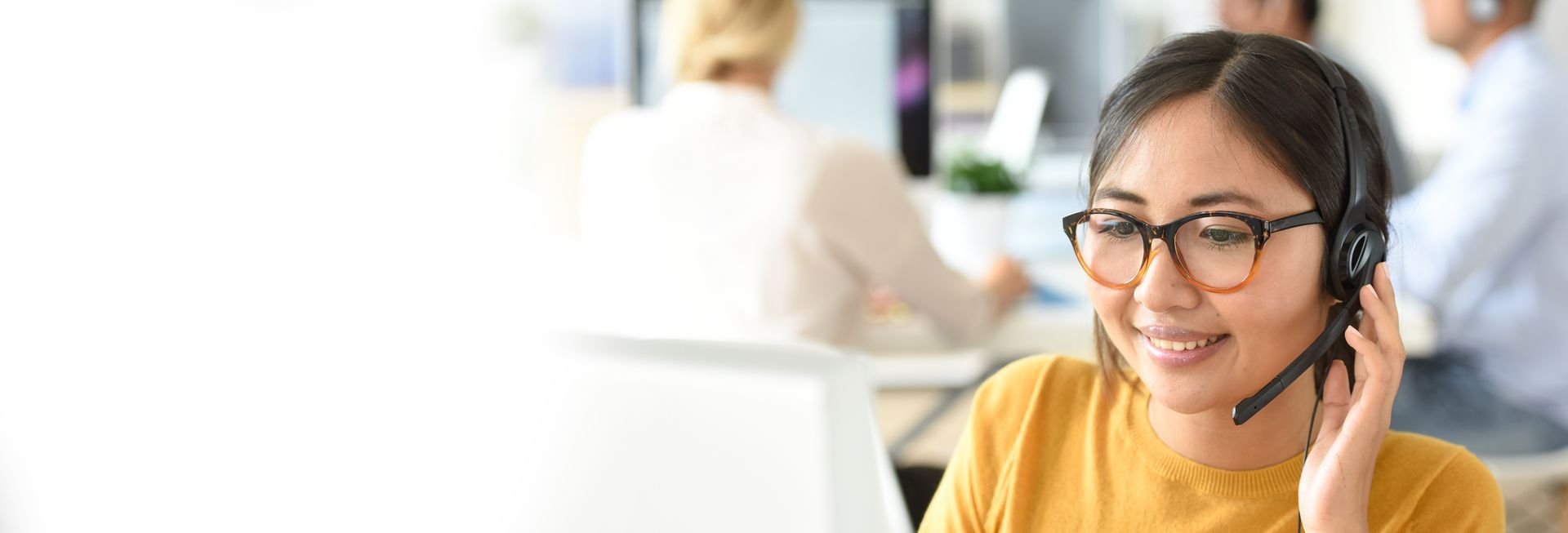 A woman wearing glasses and a headset is talking on a phone in a call center.