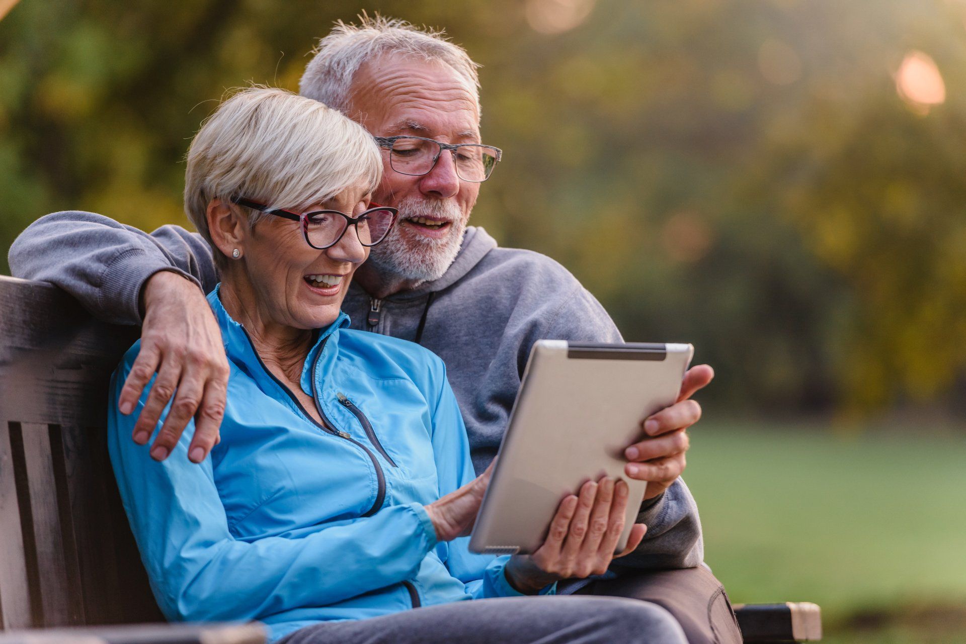 An elderly couple is sitting on a bench looking at a tablet.