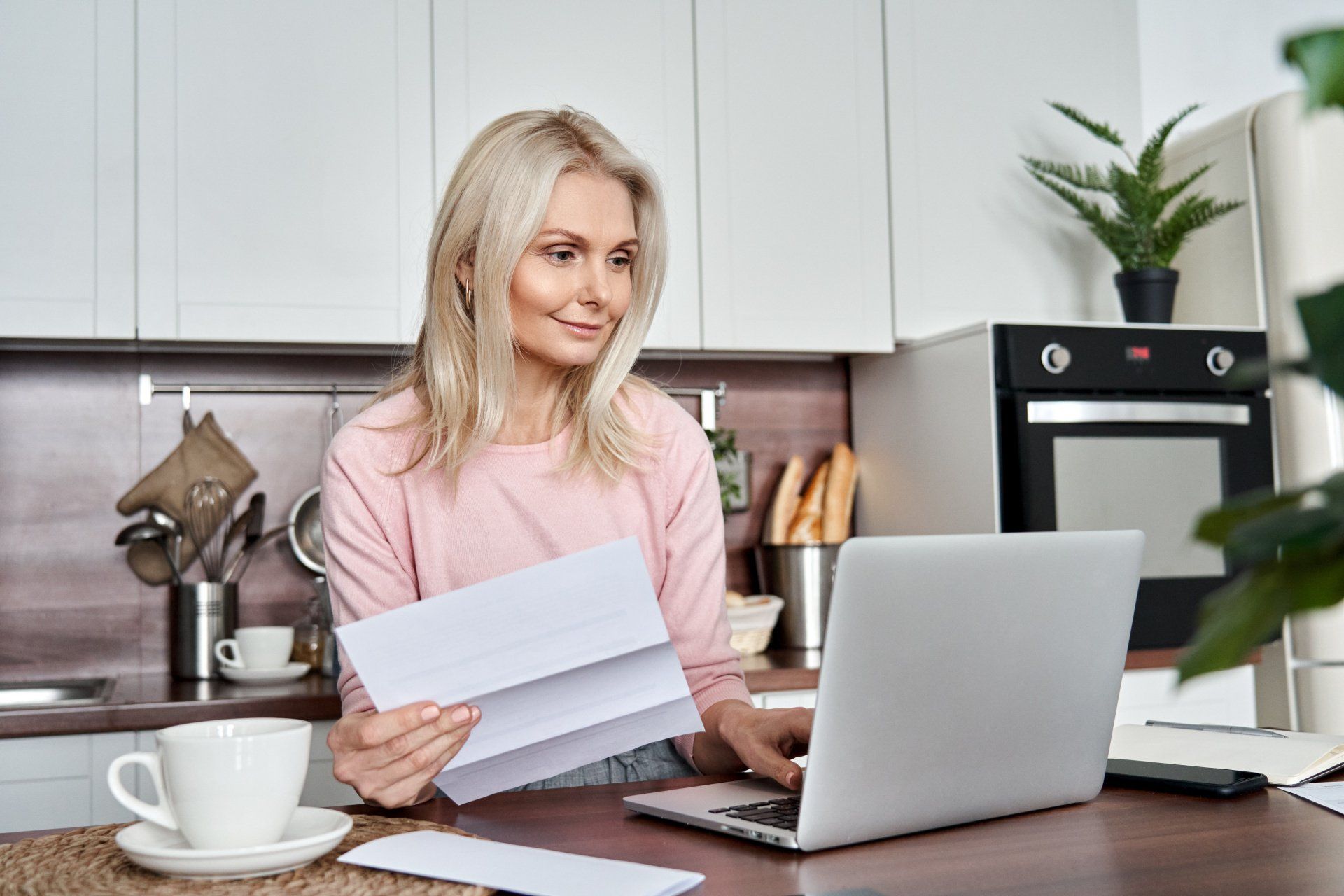 A woman is sitting at a table with a laptop and holding a piece of paper.