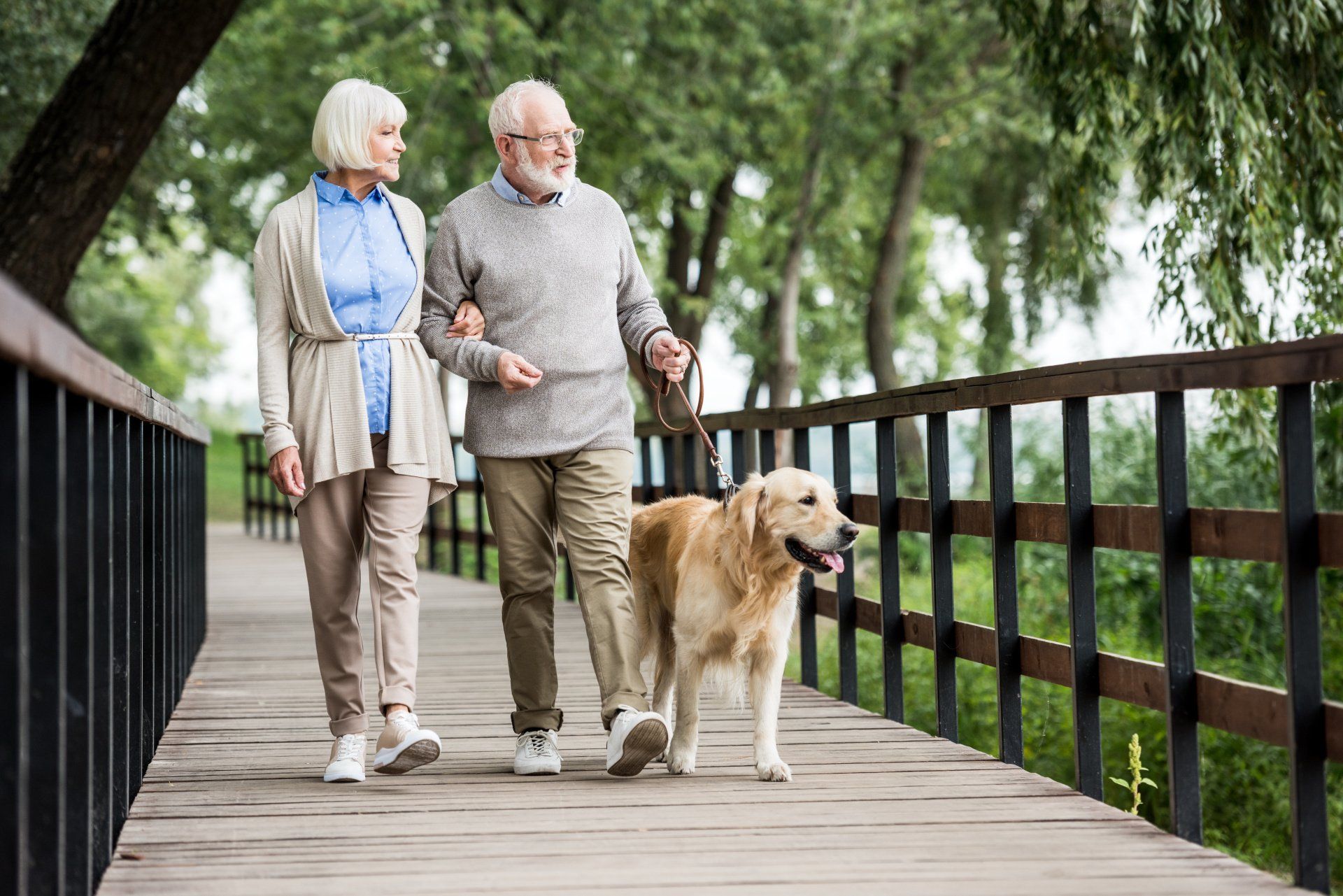 An elderly couple is walking a dog on a leash on a wooden bridge.