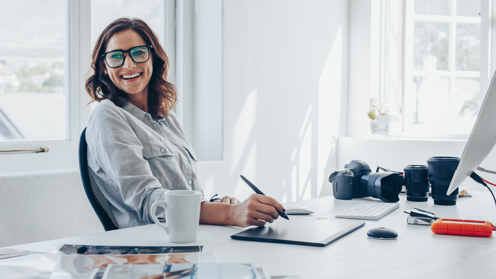 Woman smiling at desk with computer, cameras, and drawing tablet.
