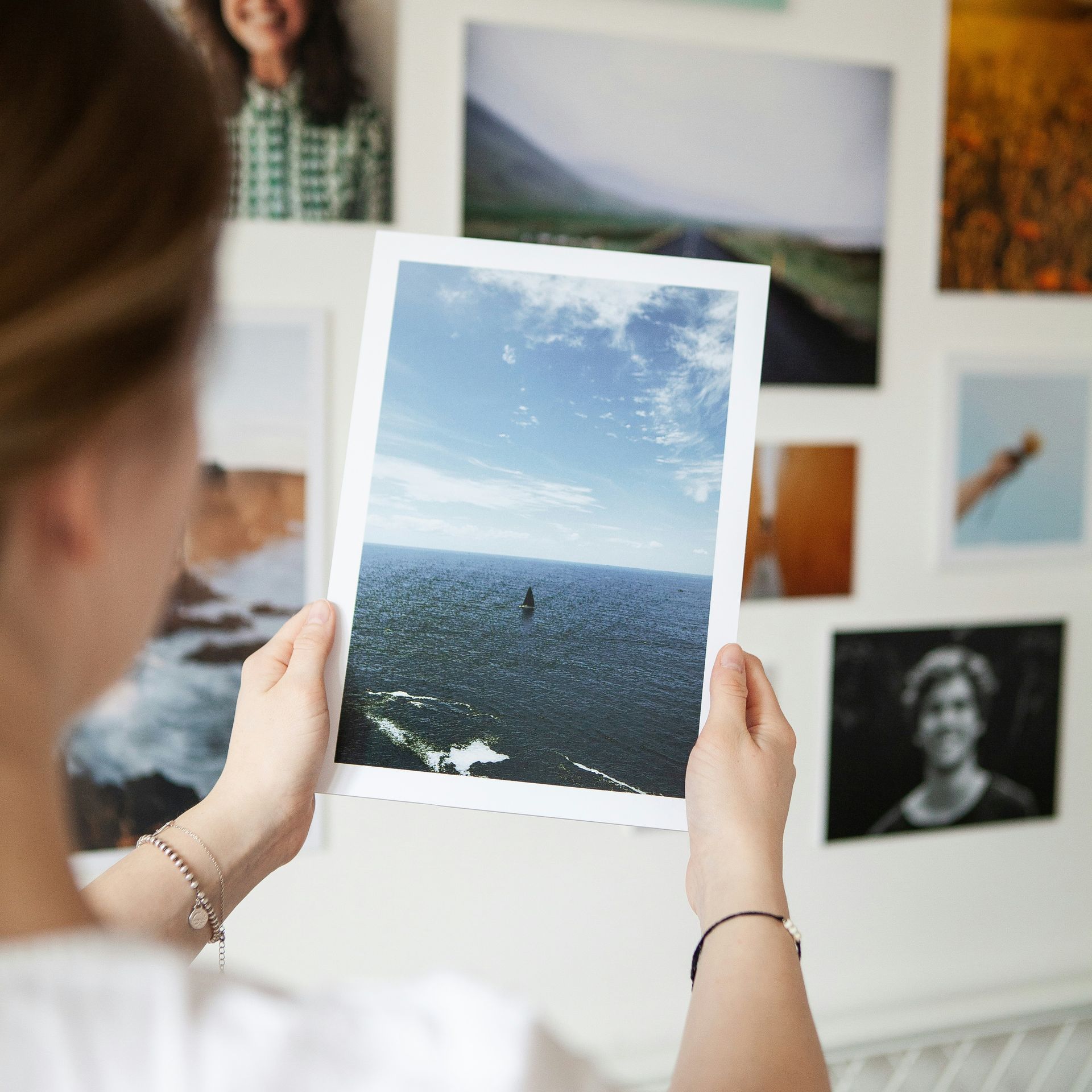 Woman holding a printed photo of a seascape, admiring it in front of a wall with other framed pictures.