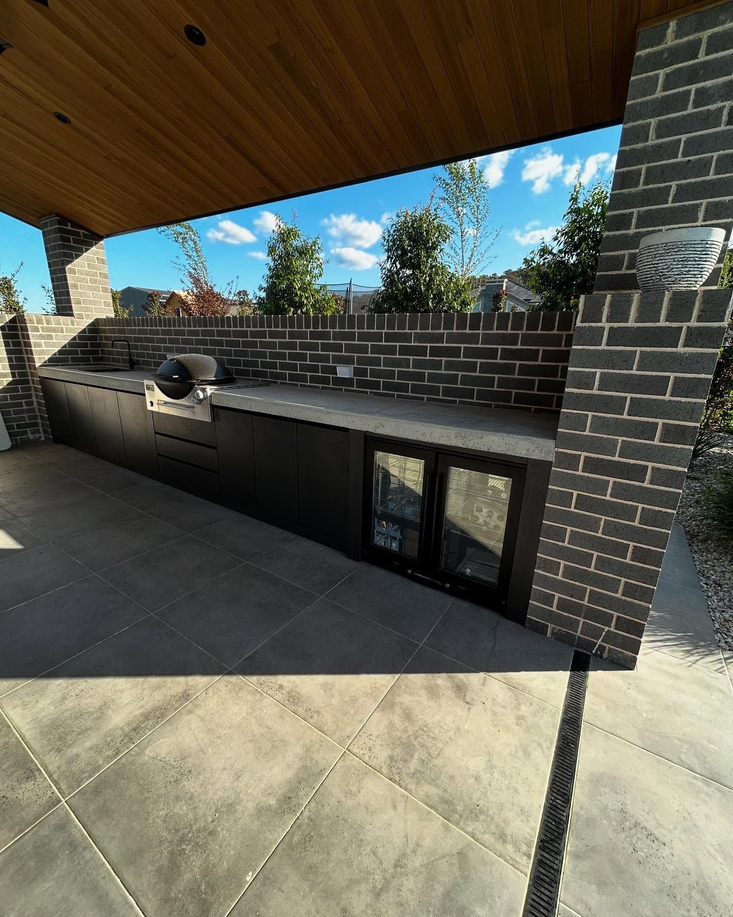 Outdoor Kitchen With Dark Cabinets, Brick Accents, Grill, And Refrigerator — Protech Renovations & Joinery in Camden, NSW