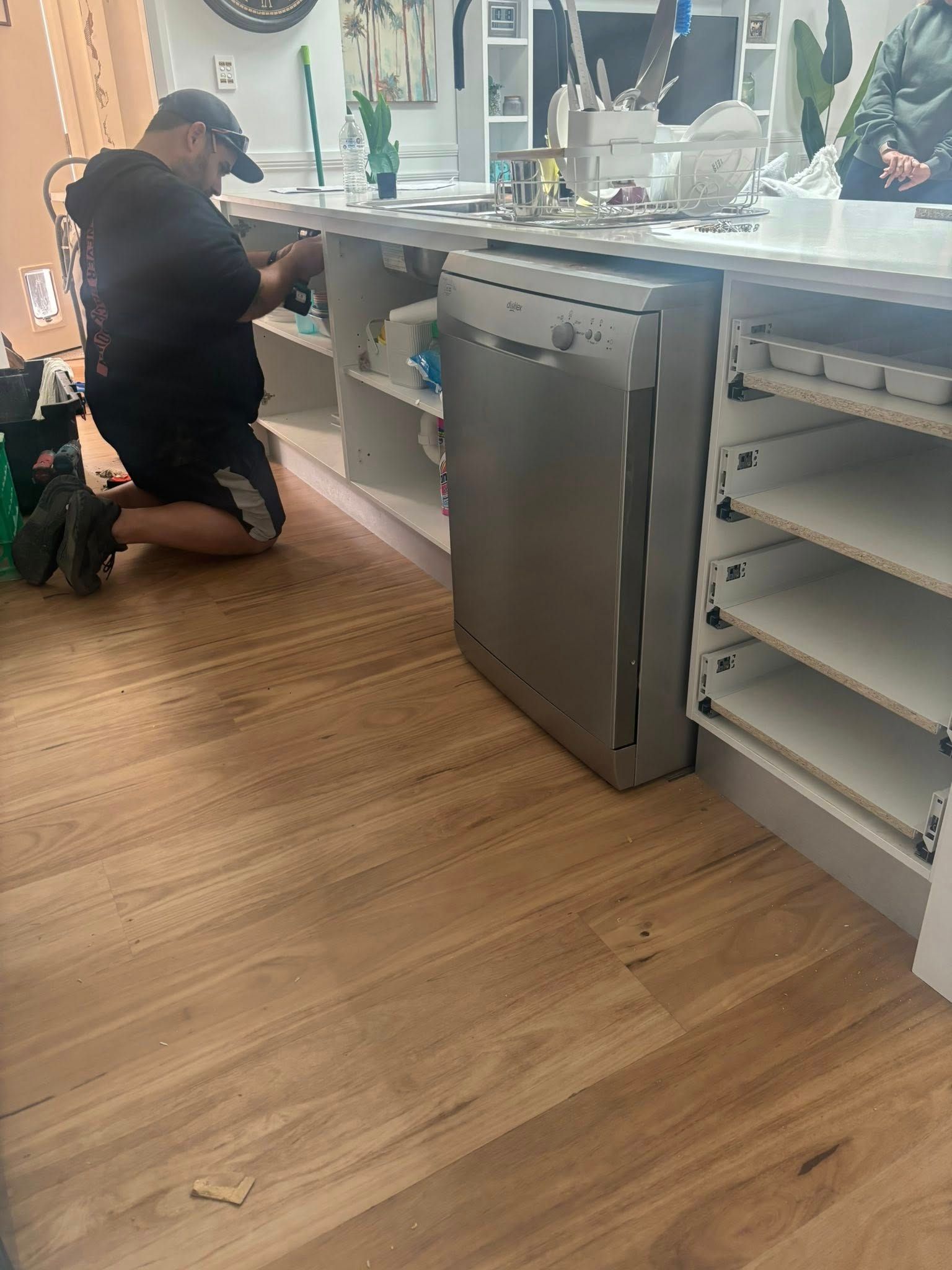 Man installing a dishwasher in a kitchen with white cabinets and wood flooring — Protech Renovations & Joinery in Gregory Hills, NSW