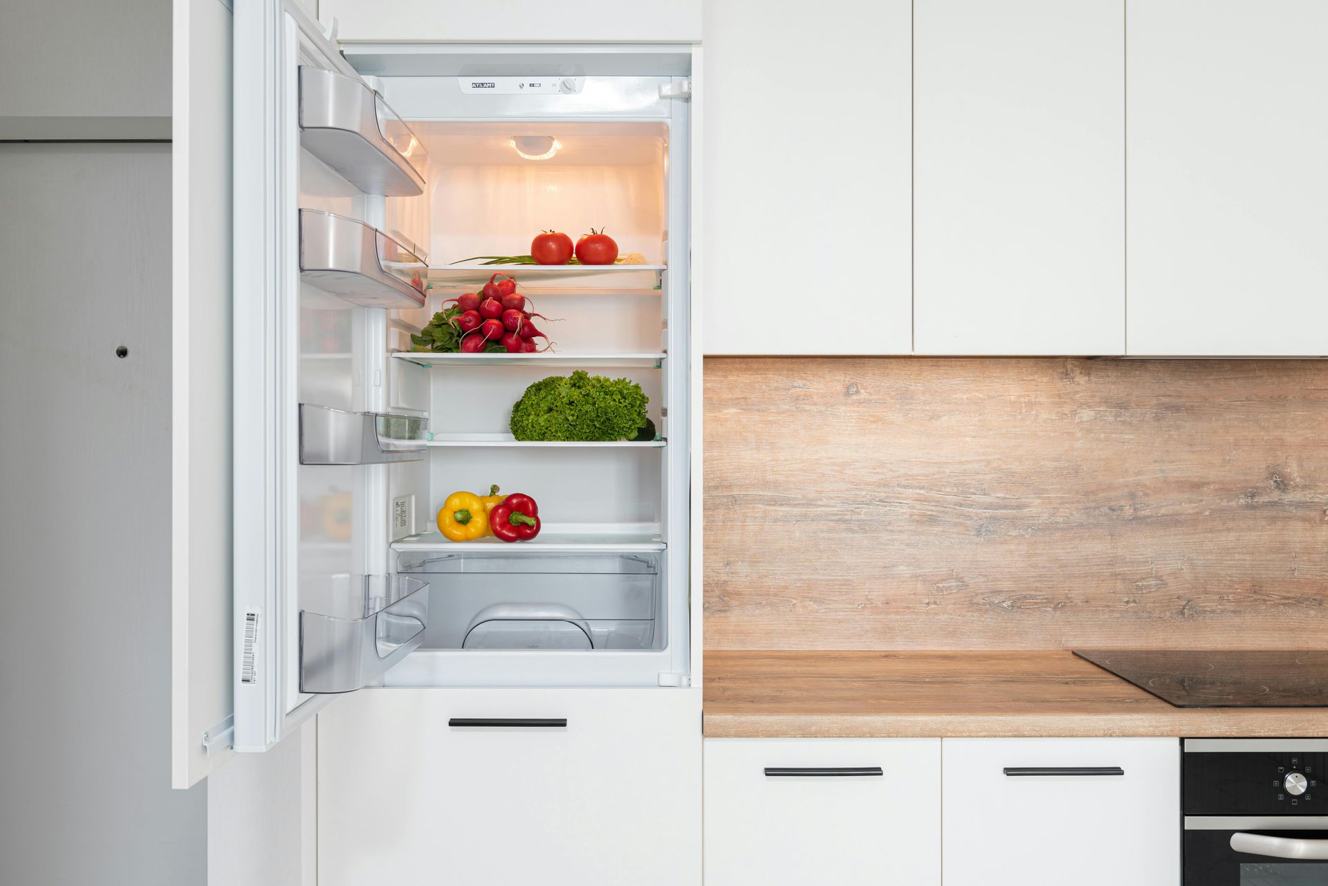 Open white refrigerator in modern kitchen showing fresh vegetables and food items stored on shelves,