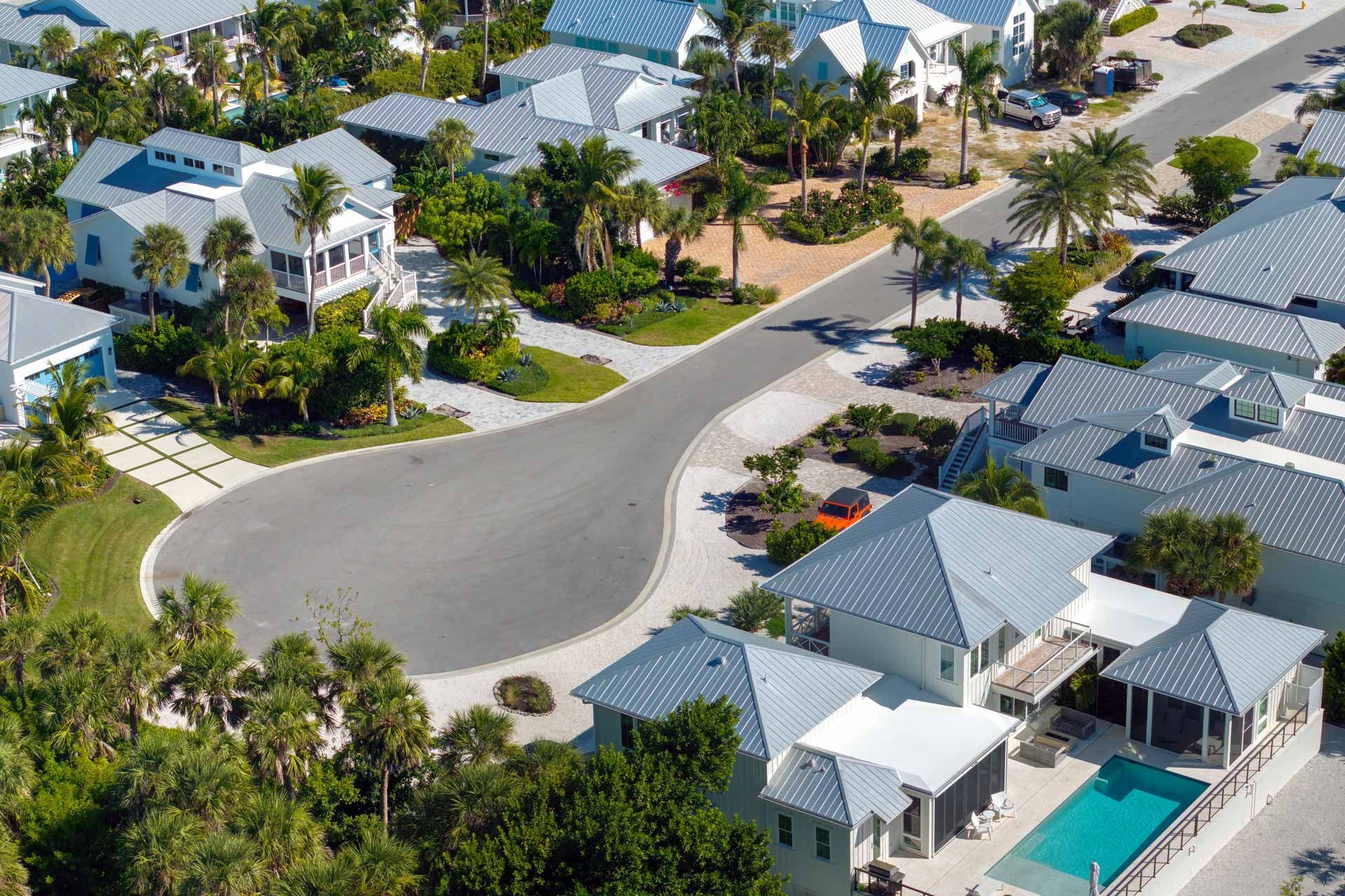 Aerial view of a coastal neighborhood with light-colored houses, blue pools, and a curved road.