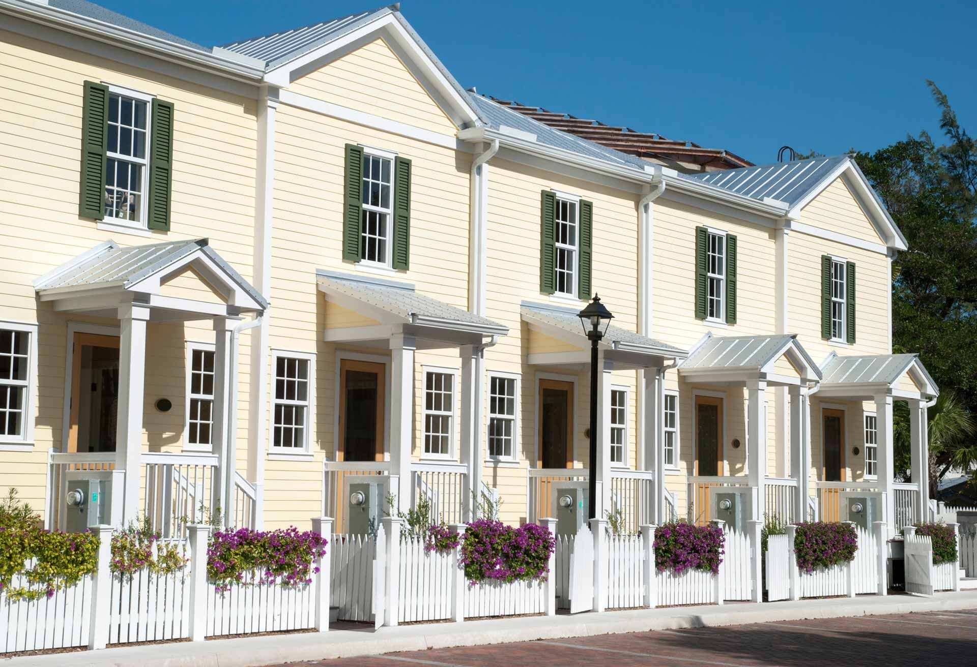 Row of yellow houses with white trim, green shutters, and a white picket fence under a blue sky.
