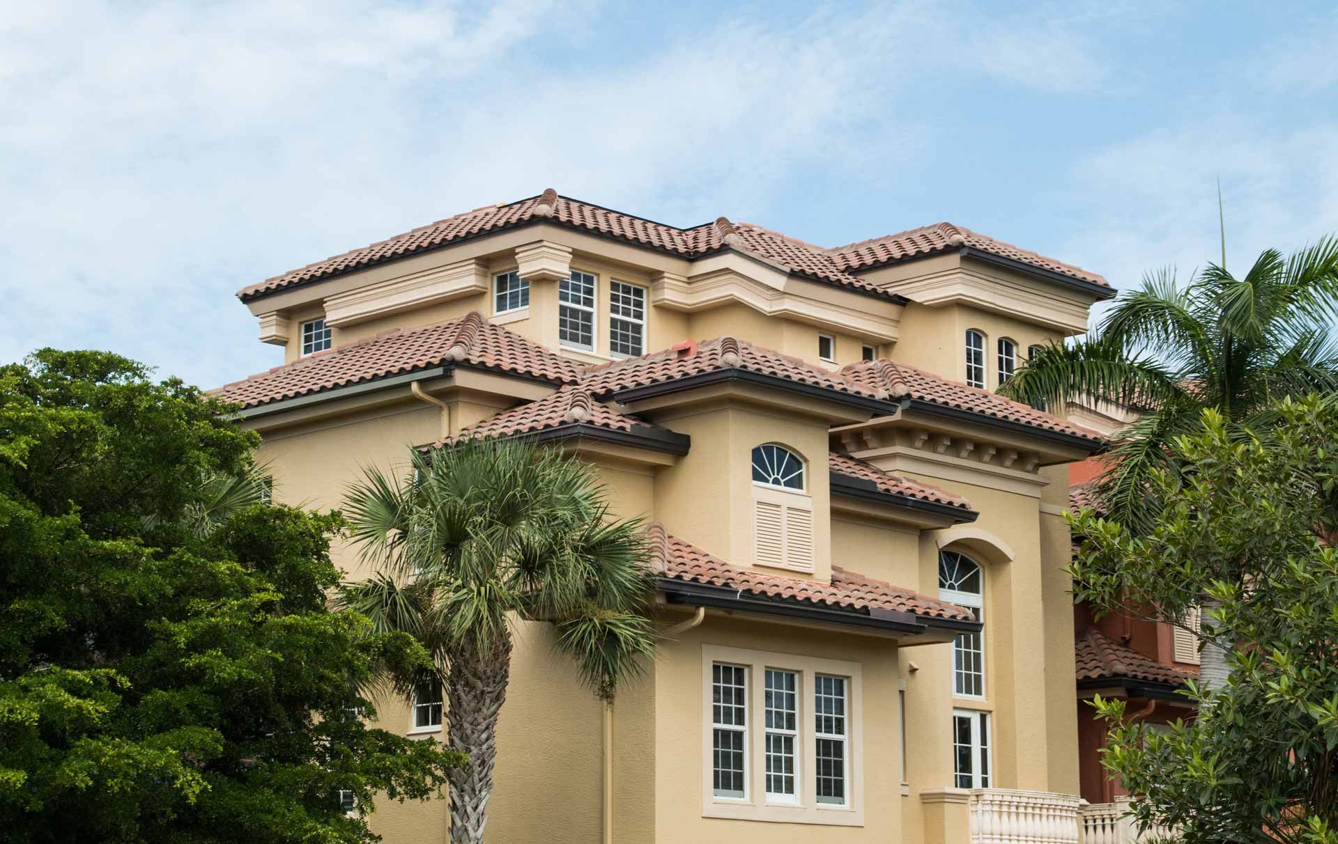 Beige stucco mansion with terracotta roof, framed by green trees and blue sky.
