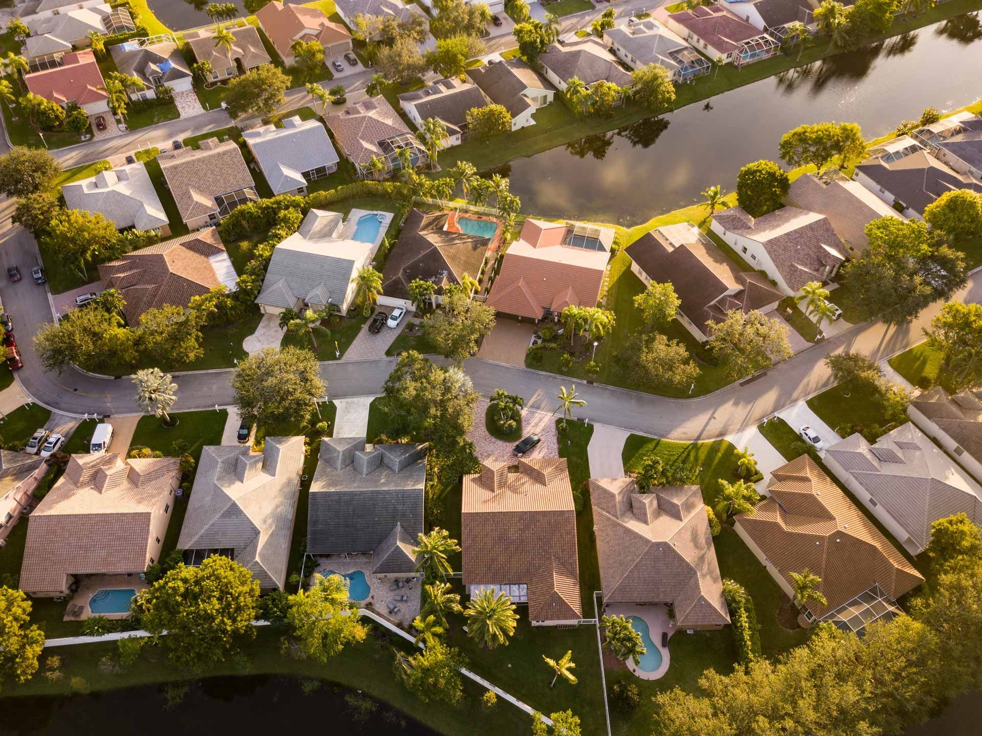 Aerial view of a suburban neighborhood with houses, streets, trees, and a lake reflecting sunlight.