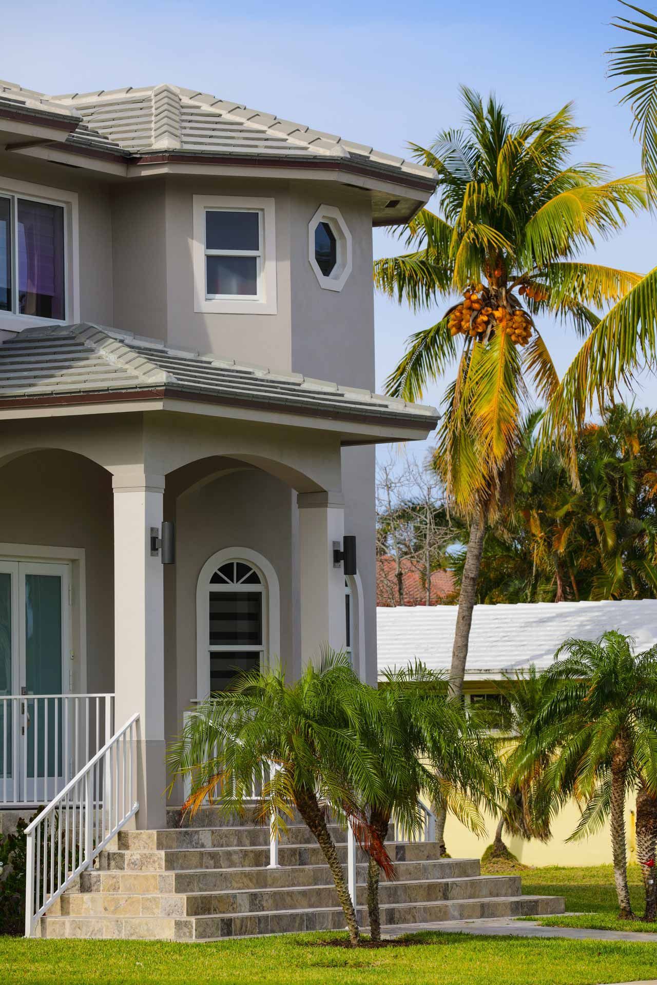 Gray stucco house with palm trees, steps, and windows under a blue sky.