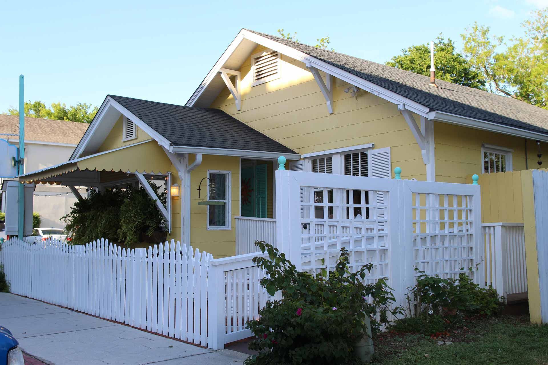 Yellow cottage with white picket fence and trellis, under a blue sky.