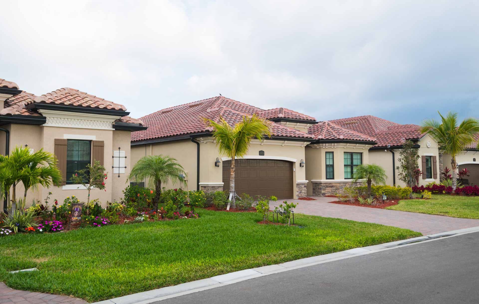 Houses with brown tile roofs, tan stucco walls, and palm trees. Driveway and green lawn in front.