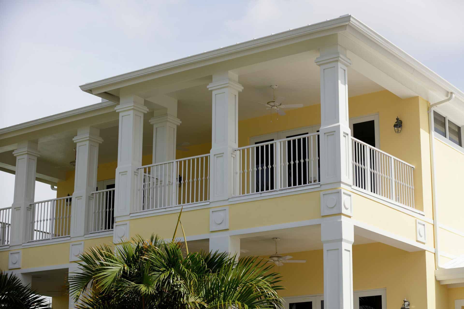 Two-story yellow house with white columns and railings, balconies on both floors. Palm trees in front.