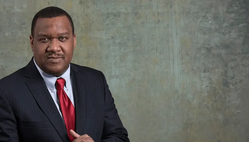 A man in a suit and red tie is standing in front of a gray wall.