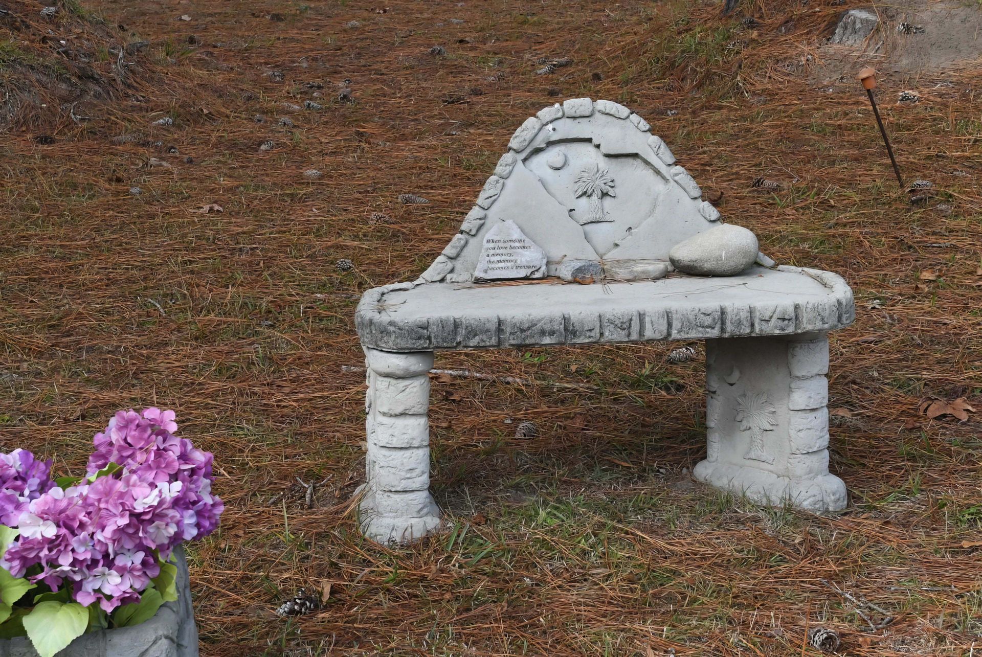 A stone bench with a heart on it sits in the grass next to a pot of purple flowers.