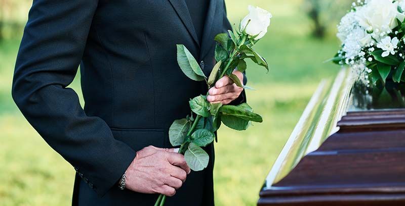 A man in a suit is holding a white rose in front of a coffin.