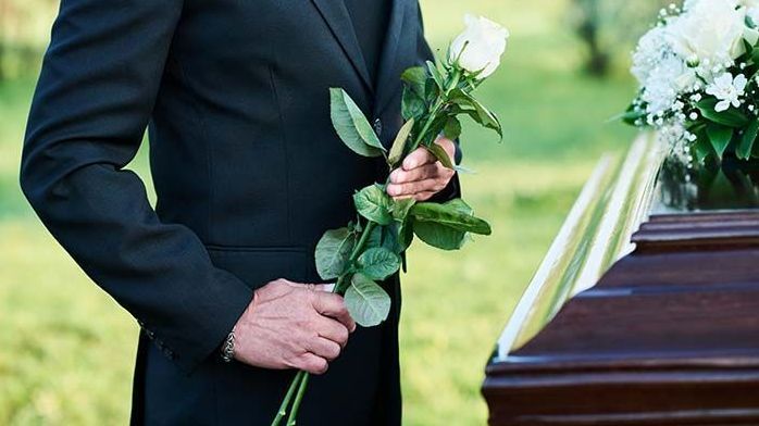 A man in a suit is holding a white rose in front of a coffin.