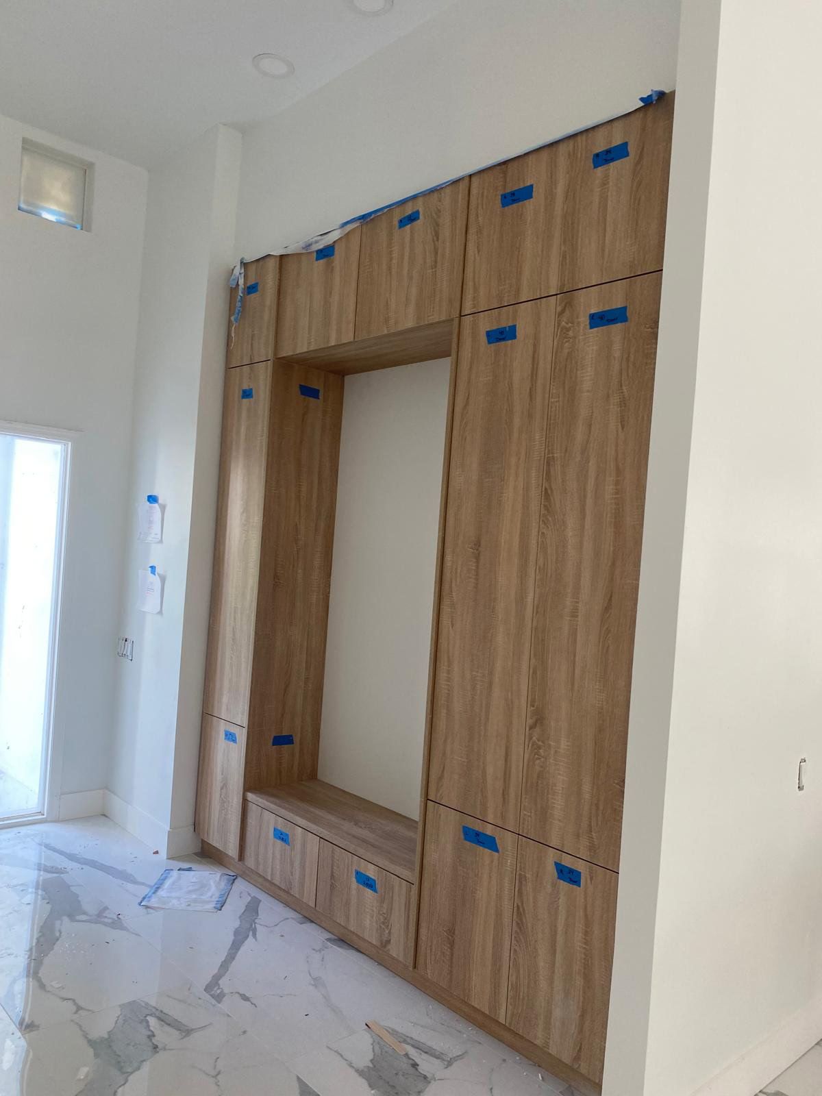 Built-in wooden cabinet unit with central open space and bench. Blue tape marks unfinished project in a bright room.